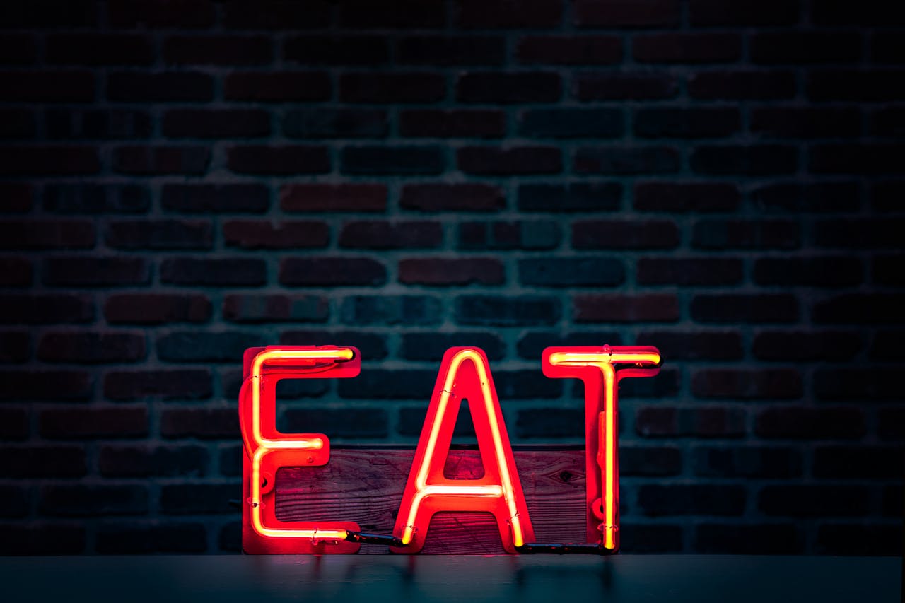 about-us-img Vibrant neon 'EAT' sign in red glowing boldly against a dark brick wall background.